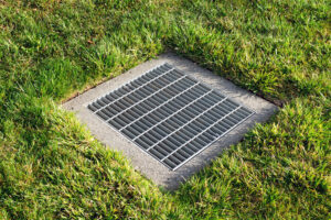 Catch basin square drainage grate over storm water drain surrounded by green grass.